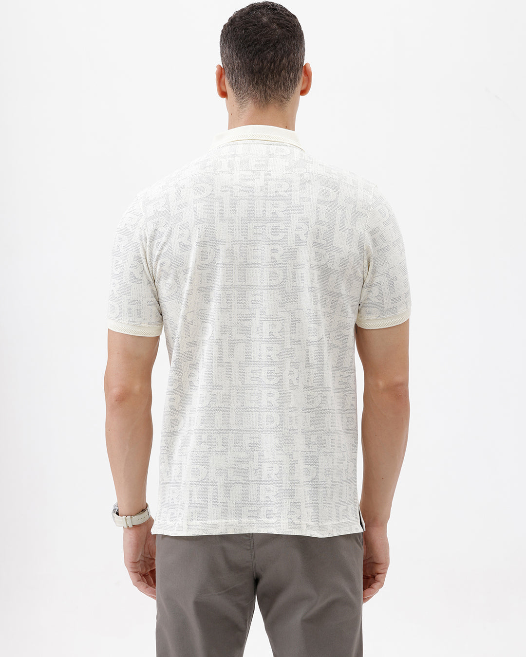 A man wearing a short-sleeve polo shirt with a subtle repeating monogram pattern in cream and light gray, paired with gray trousers, photographed against a plain white studio background.