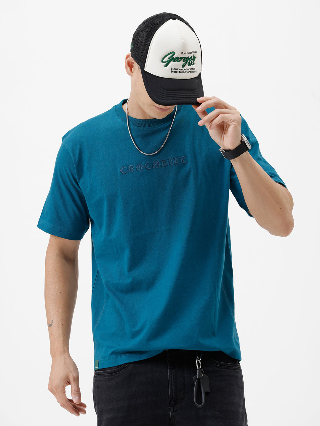 A man wearing a teal blue short-sleeve t-shirt with a subtle tonal 'GREATNESS' text graphic across the chest, paired with black pants, photographed against a clean white studio background.
