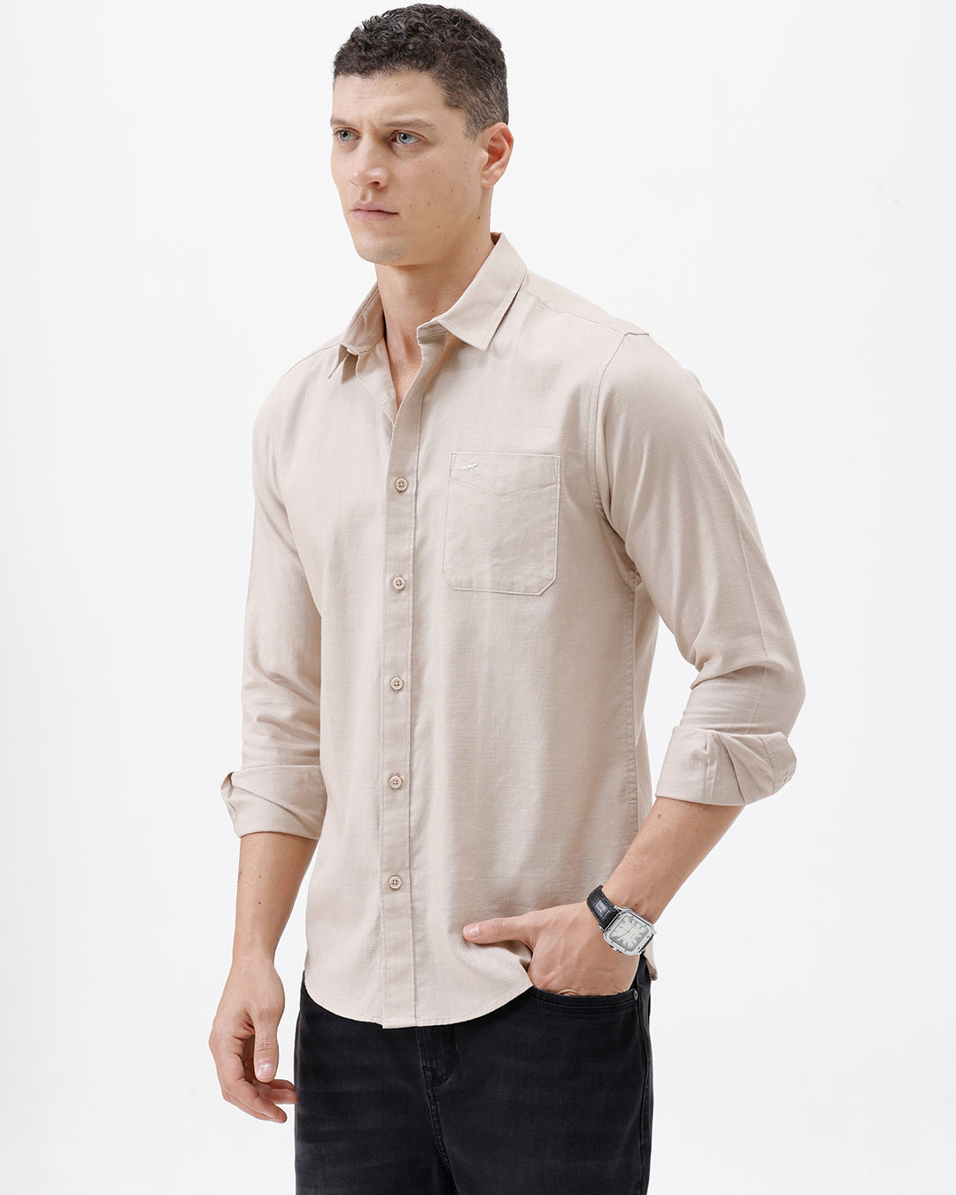 A man wearing a light beige long-sleeve button-up shirt with a chest pocket, paired with dark jeans, standing against a plain white studio background.
