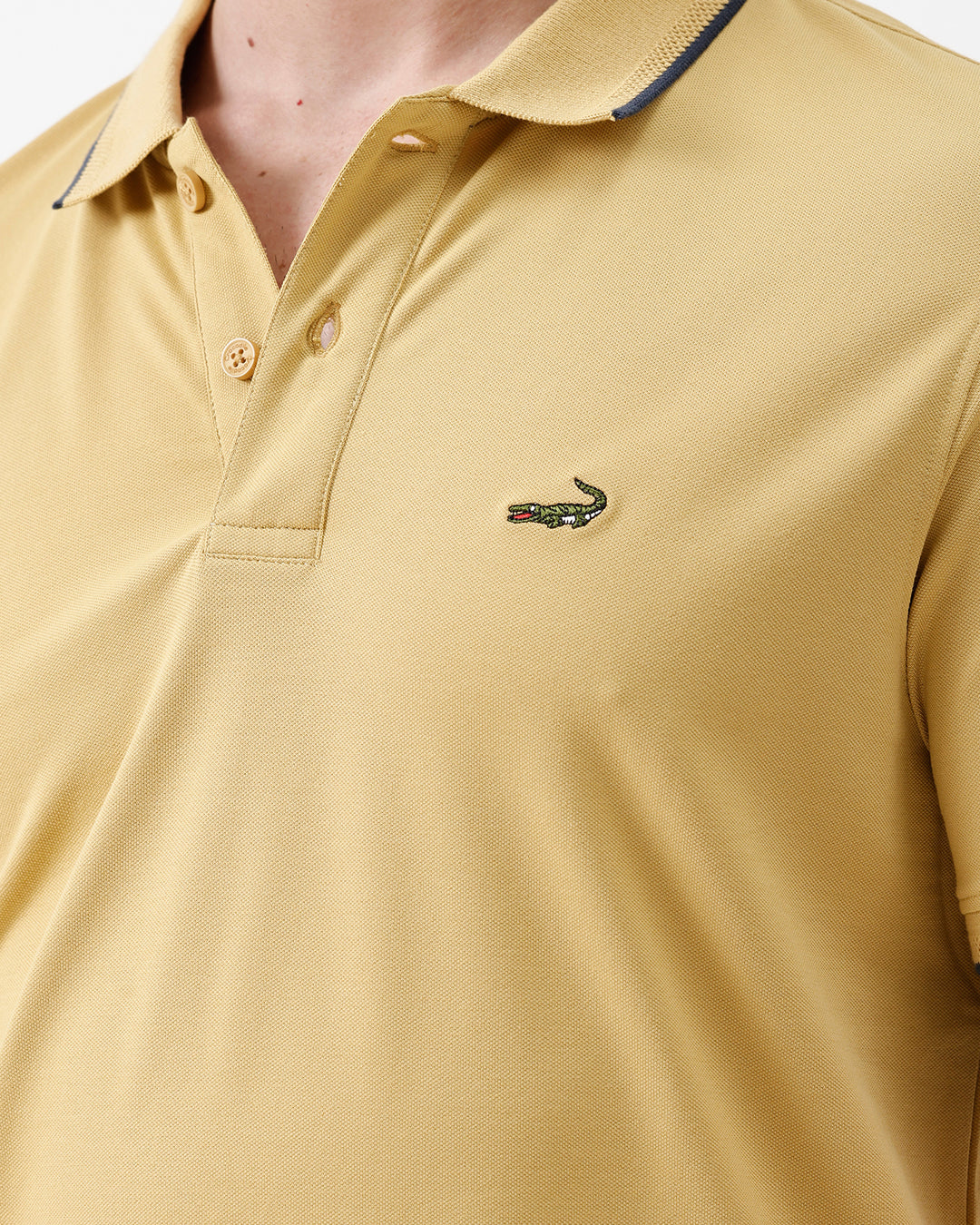 A man wearing a light yellow Lacoste polo shirt with dark piping on the collar and sleeve cuffs, paired with beige trousers, against a plain white studio background.