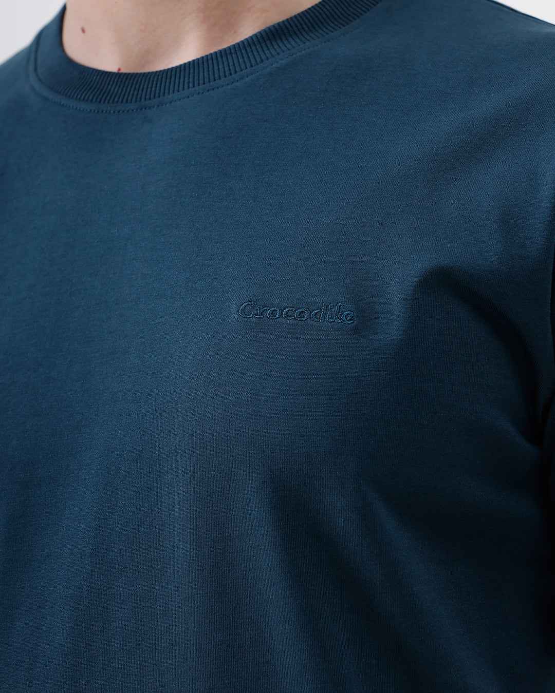 A man wearing a dark navy blue crew-neck t-shirt and black jeans, standing against a light gray studio background.
