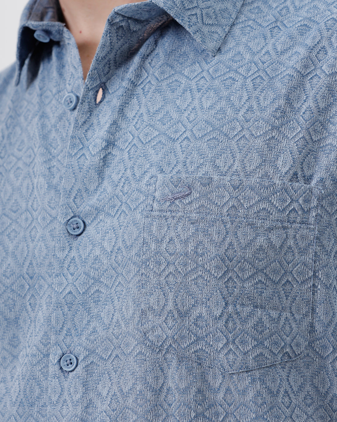 A man wearing a short-sleeve button-up shirt with a fine blue and white geometric pattern, paired with light beige trousers, standing against a plain light gray background.