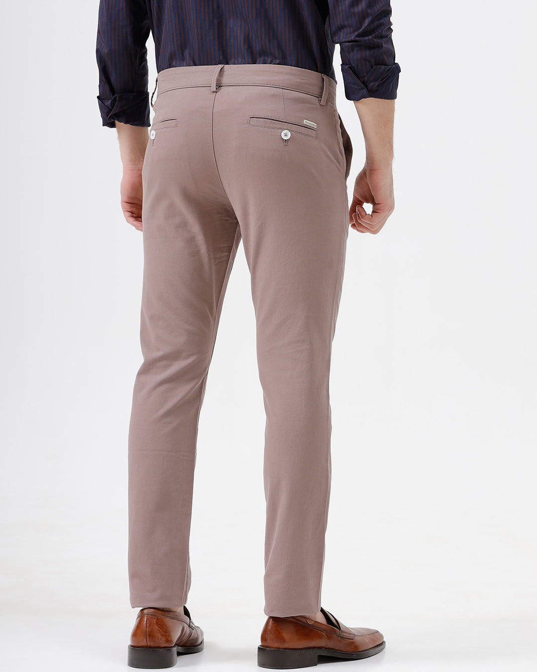 A person wearing slim-fit taupe trousers, a navy blue striped button-down shirt, and brown leather loafers, standing against a plain white studio background.