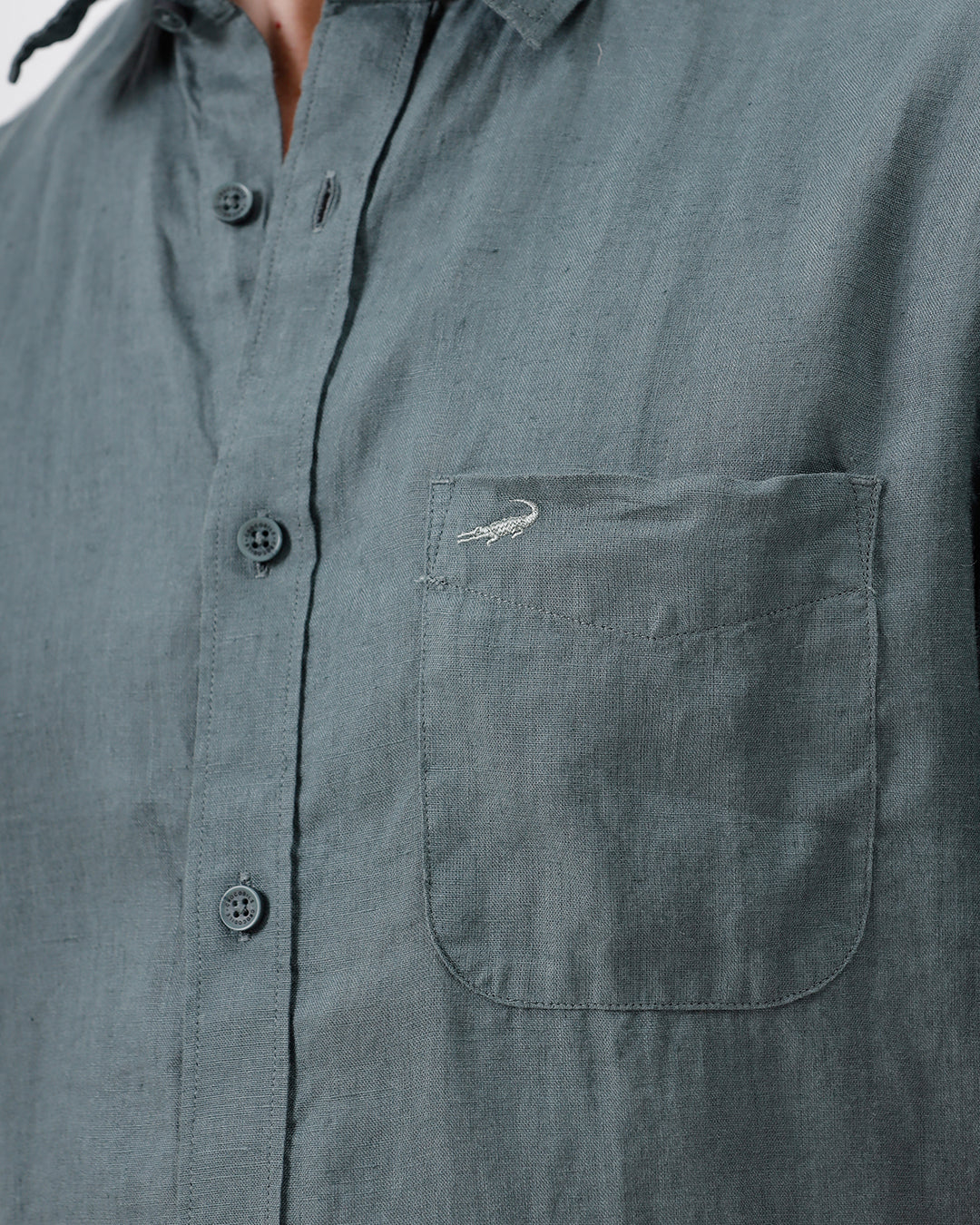 A man wearing a short-sleeve button-up shirt in a muted slate green color, paired with light beige trousers, standing against a plain light gray background.