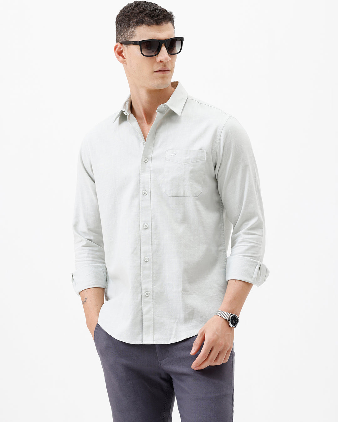 A man wearing a light sage green long-sleeve button-down shirt with rolled-up sleeves and dark gray trousers, standing against a plain white studio background.