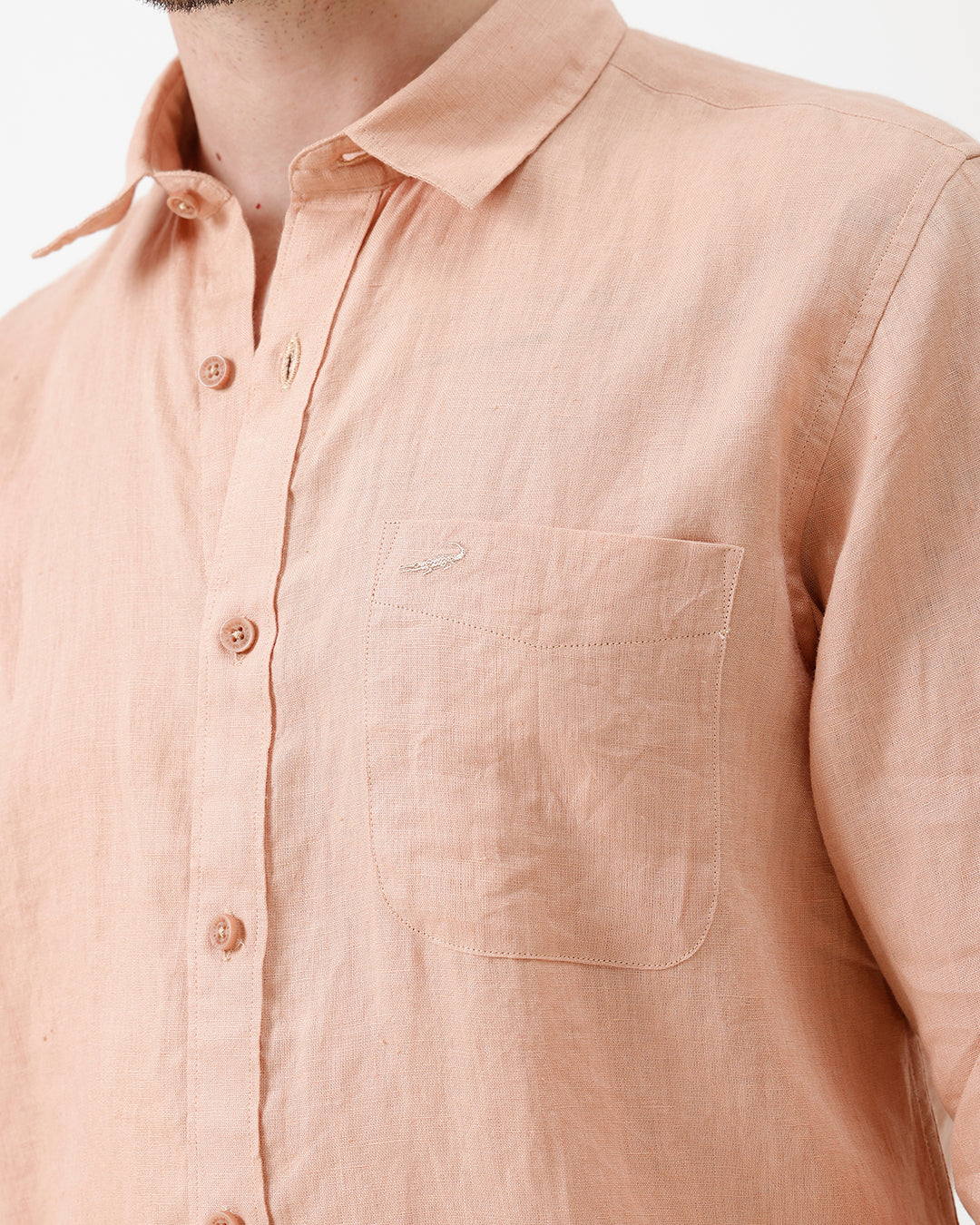 A man wearing a light peach-colored linen button-down shirt with dark jeans, standing against a plain white studio background.