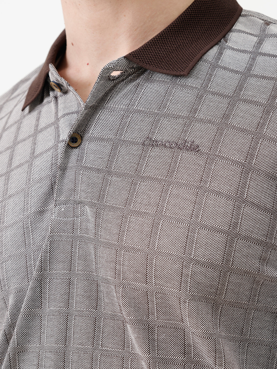 A man wearing a gray short-sleeve polo shirt with a subtle grid pattern and dark brown collar and sleeve trim, paired with dark blue jeans, against a plain white studio background.