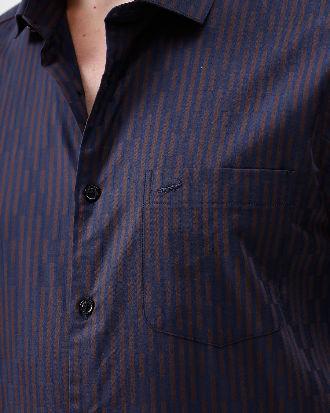 A man wearing a dark navy blue button-down shirt with thin vertical red stripes and light brown trousers, standing against a plain white studio background.