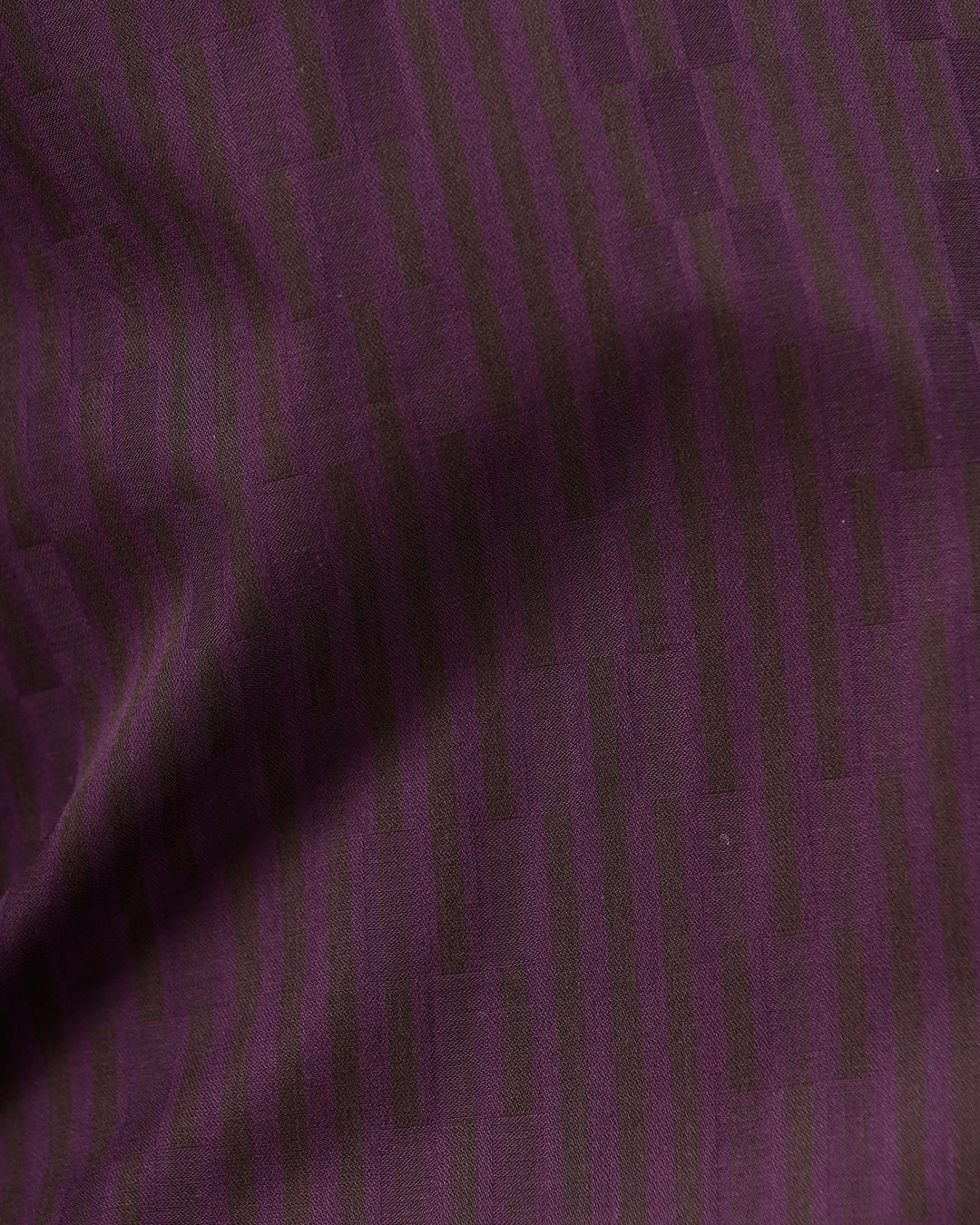 A man wearing a dark purple long-sleeve button-down shirt with subtle vertical stripes and dark navy trousers, standing against a light gray background.