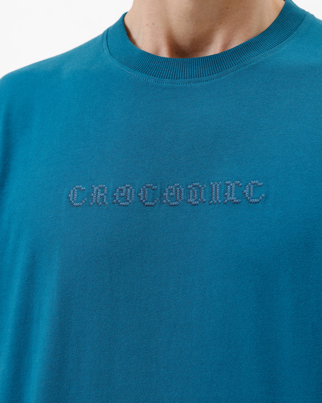 A man wearing a teal blue short-sleeve t-shirt with a subtle tonal 'GREATNESS' text graphic across the chest, paired with black pants, photographed against a clean white studio background.