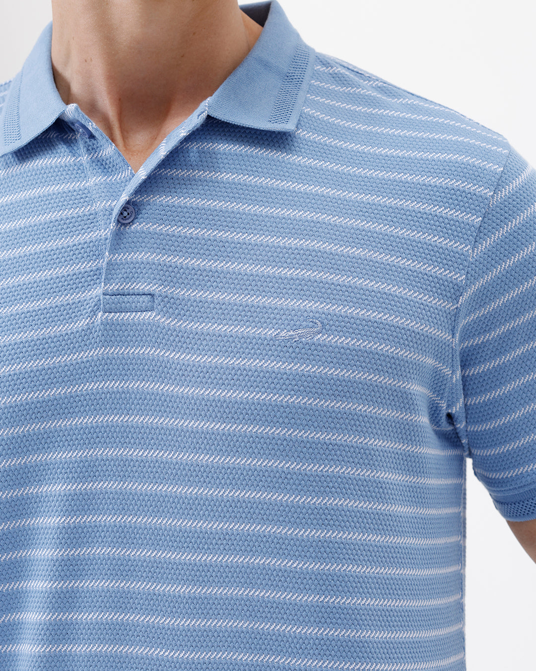 A man wearing a light blue and white striped short-sleeve polo shirt with dark blue jeans, standing against a plain light gray background.