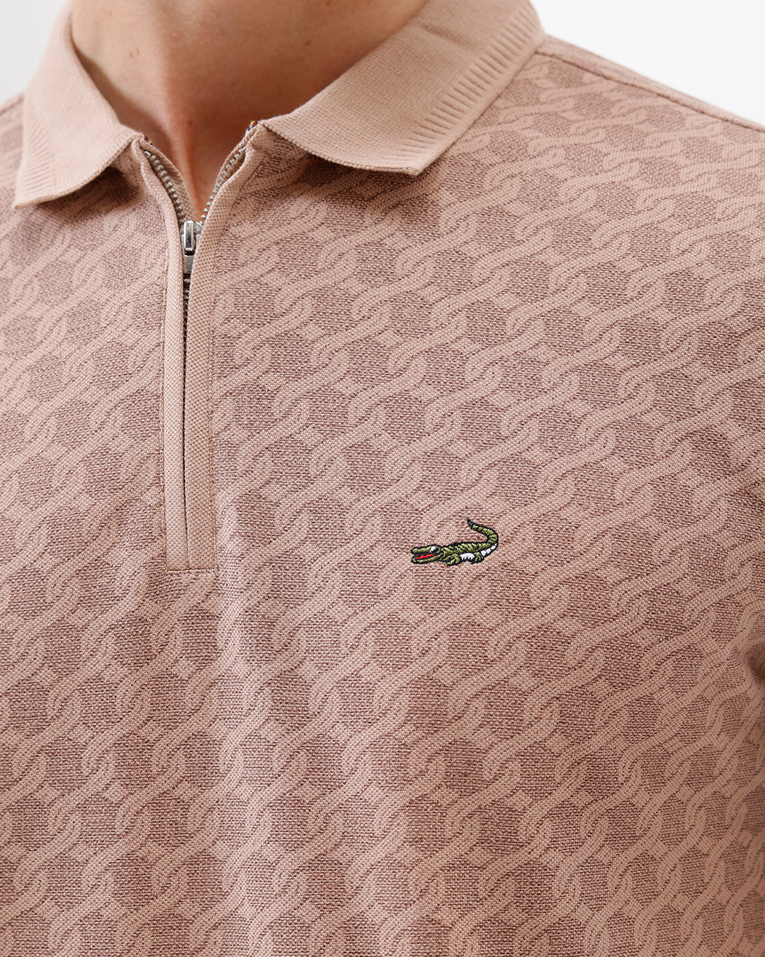 A man wearing a short-sleeve polo shirt with a muted brown and beige geometric pattern, paired with dark gray trousers, standing against a plain white studio background.