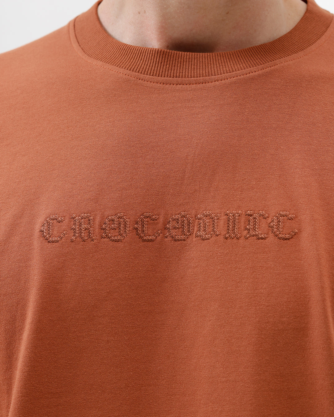 A man wearing a rust-orange crewneck t-shirt with a subtle tonal 'ELECTRIC' logo across the chest, paired with black pants, photographed against a clean white studio background.
