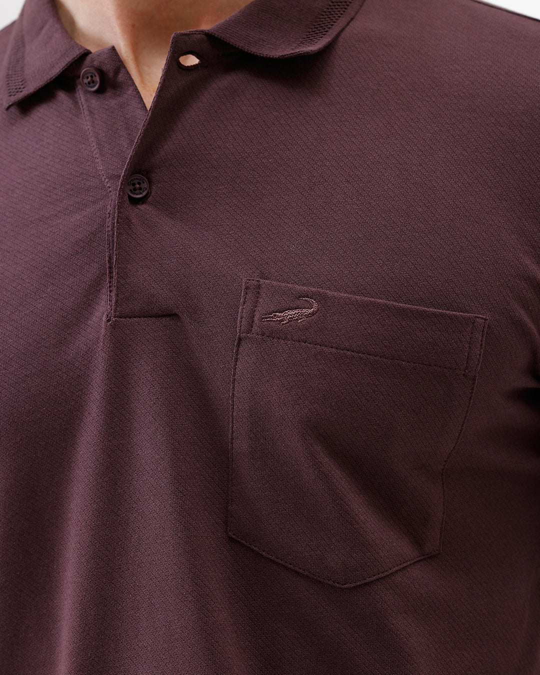 A man wearing a short-sleeve polo shirt in dark burgundy with a chest pocket, paired with gray trousers, standing against a plain light gray background.