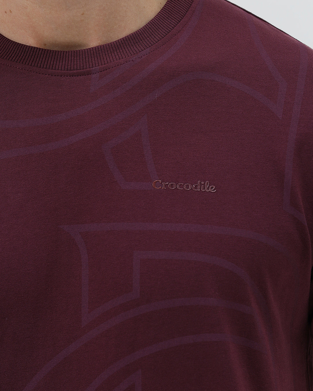 A man wearing a short-sleeve crewneck t-shirt in maroon with a subtle tonal graphic pattern, paired with dark blue jeans, against a light gray studio background.