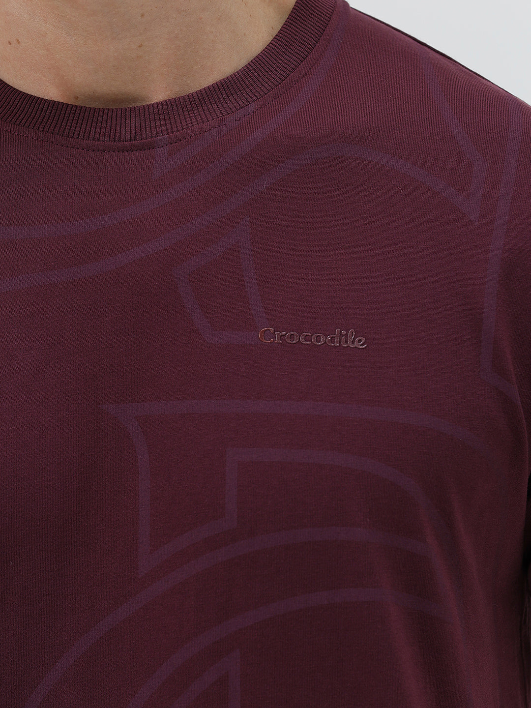 A man wearing a short-sleeve crewneck t-shirt in maroon with a subtle tonal graphic pattern, paired with dark blue jeans, against a light gray studio background.