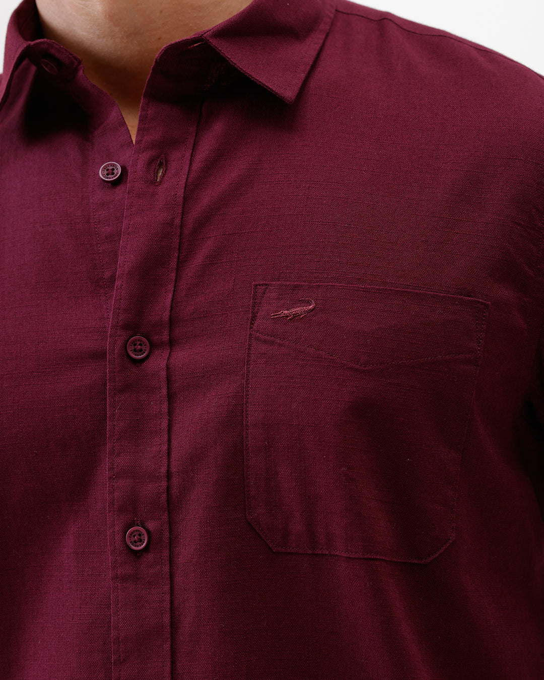A man wearing a short-sleeve button-up shirt in maroon with a chest pocket, paired with blue jeans, standing against a plain white studio background.