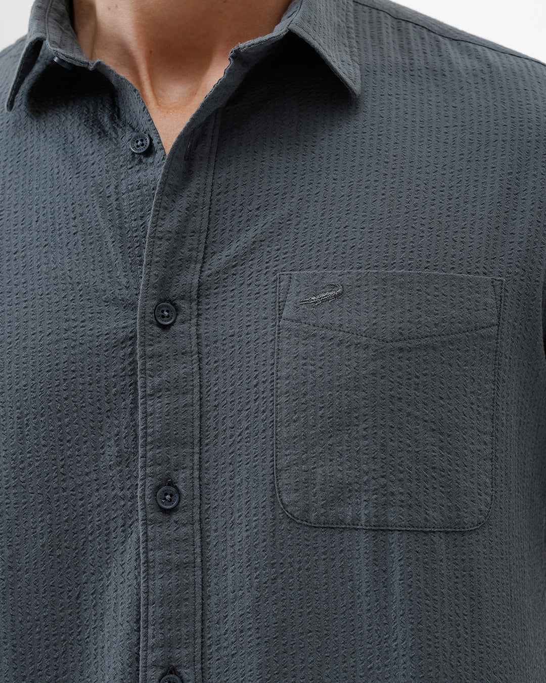 A man wearing a dark charcoal gray long-sleeve button-down shirt with a chest pocket, paired with light gray trousers, standing against a plain white studio background.