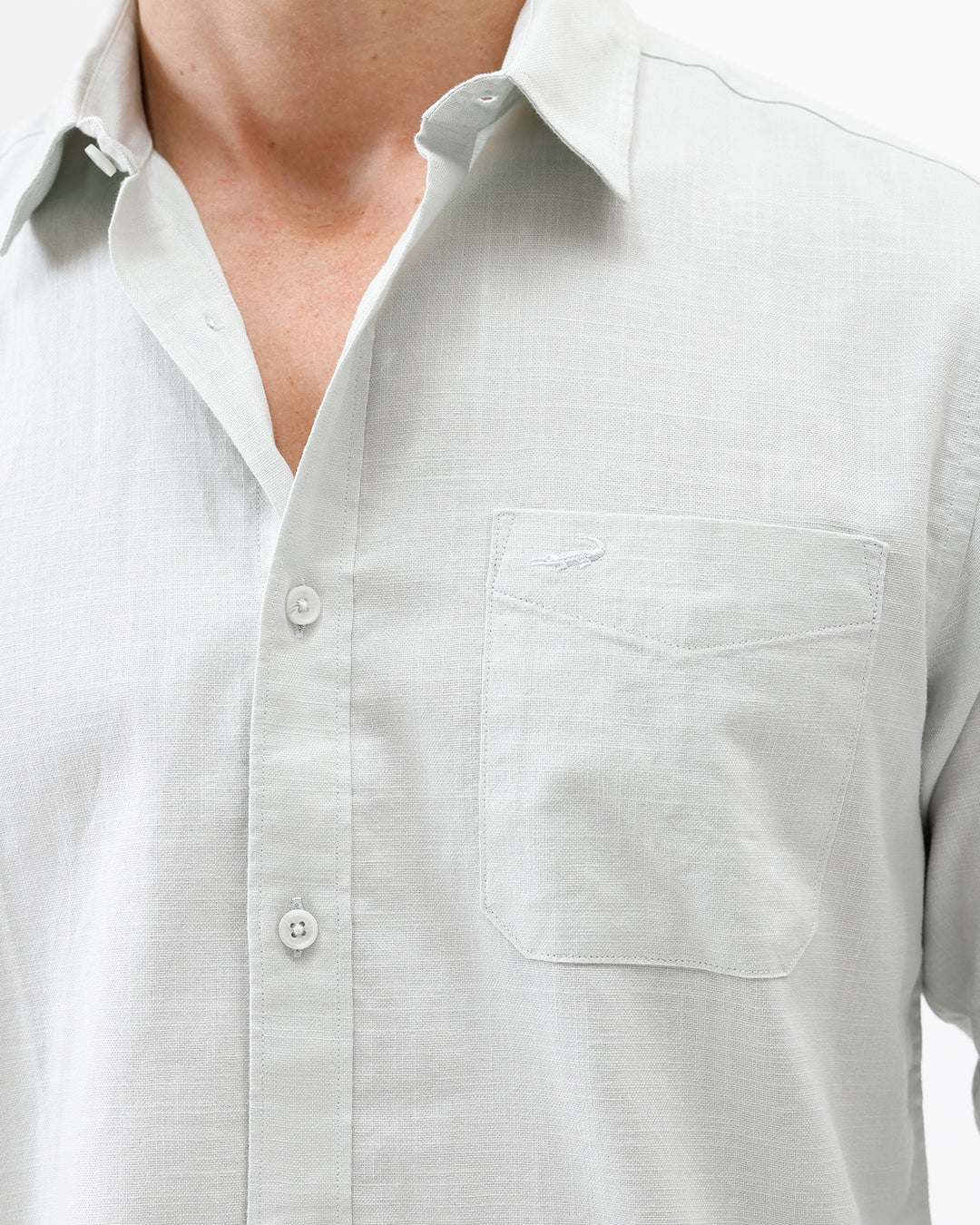 A man wearing a light sage green long-sleeve button-down shirt with rolled-up sleeves and dark gray trousers, standing against a plain white studio background.