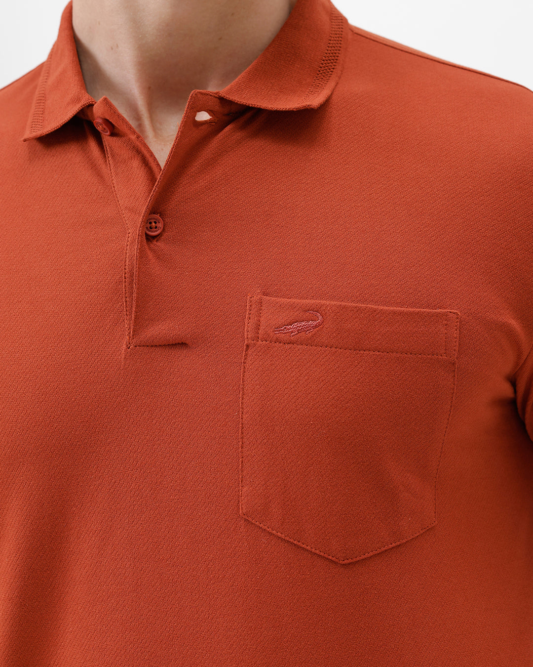A man wearing a rust-orange short-sleeve polo shirt with a chest pocket, paired with beige cargo pants, standing against a plain light gray studio background.