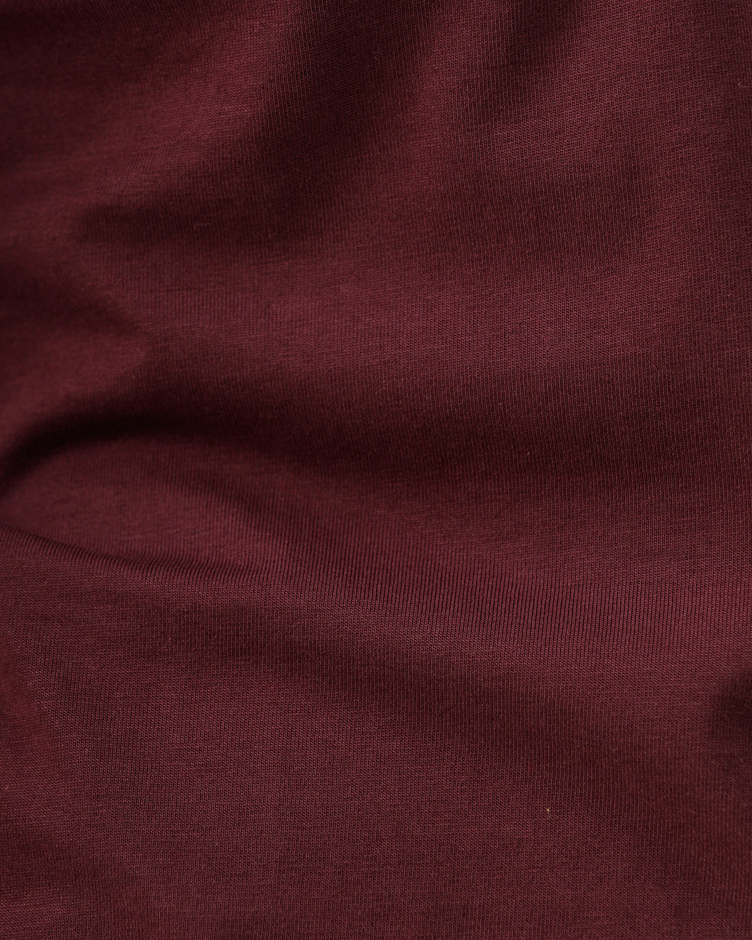 A man wearing a short-sleeve crewneck t-shirt in maroon, paired with dark jeans, standing against a plain light gray studio background.