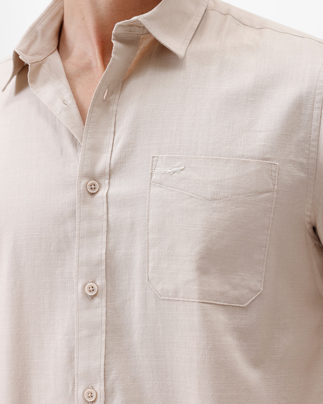A man wearing a light beige long-sleeve button-up shirt with a chest pocket, paired with dark jeans, standing against a plain white studio background.