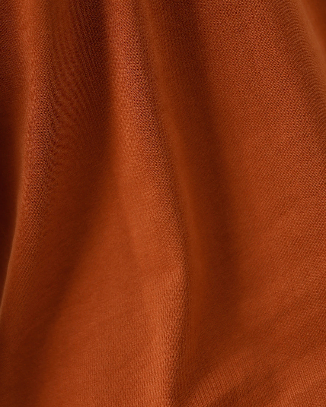 A man wearing a rust-orange crew-neck t-shirt and dark blue jeans, standing against a light gray studio background.