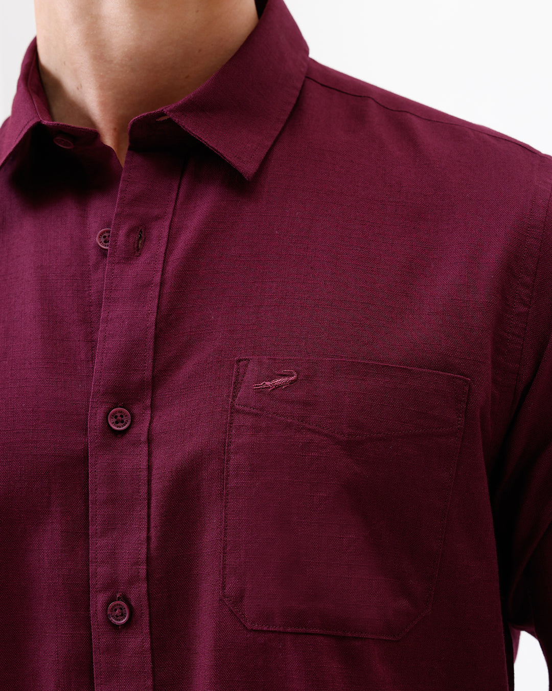 A man wearing a long-sleeve button-down shirt in deep burgundy with rolled-up sleeves and gray trousers, standing against a plain white studio background.
