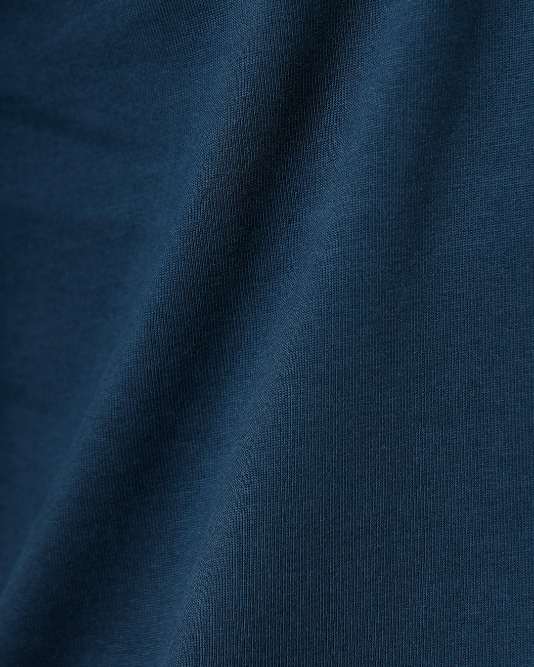 A man wearing a dark navy blue crew-neck t-shirt and black jeans, standing against a light gray studio background.
