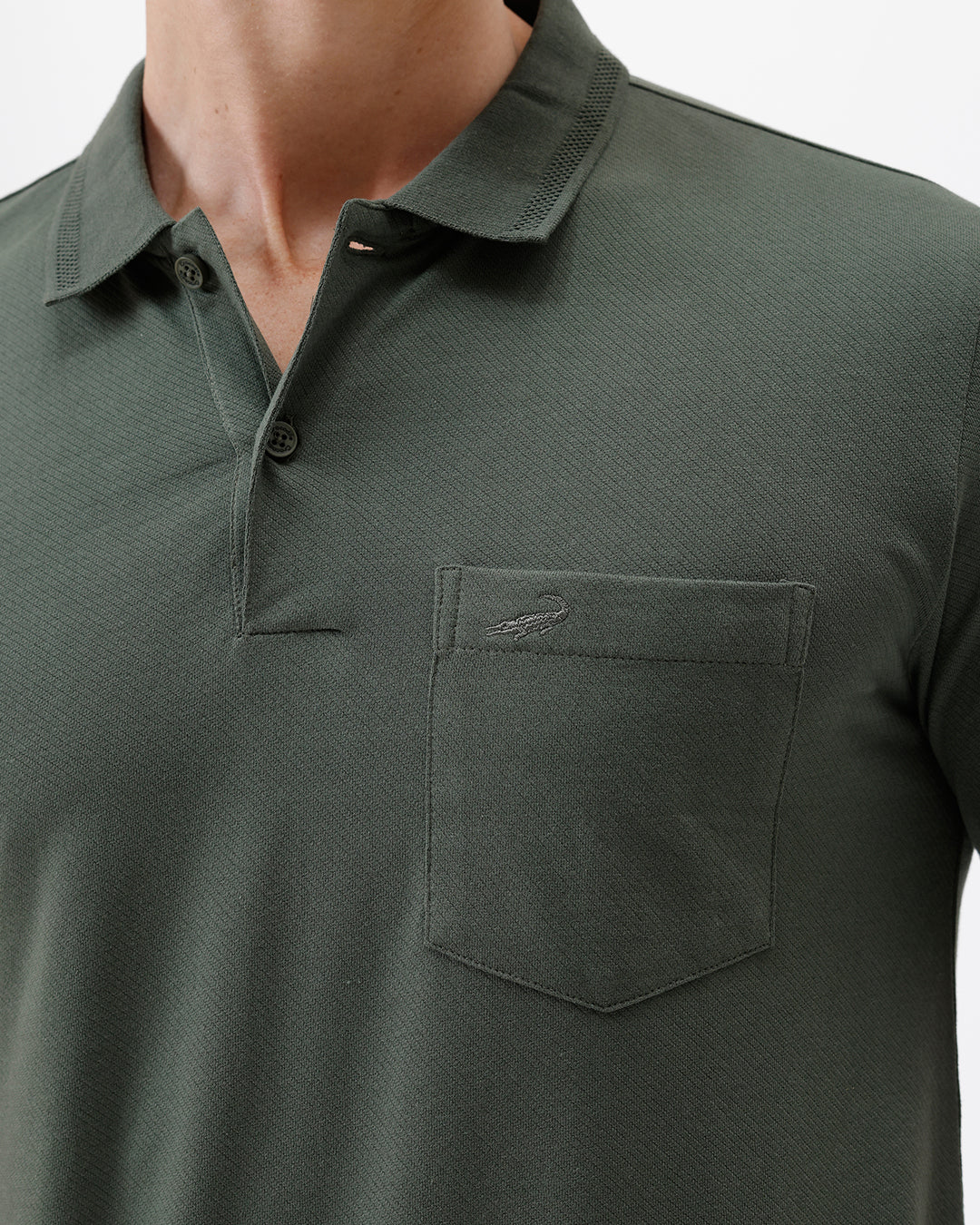 A man wearing a dark olive green short-sleeve polo shirt with a chest pocket, paired with dark jeans, standing against a plain light gray background.
