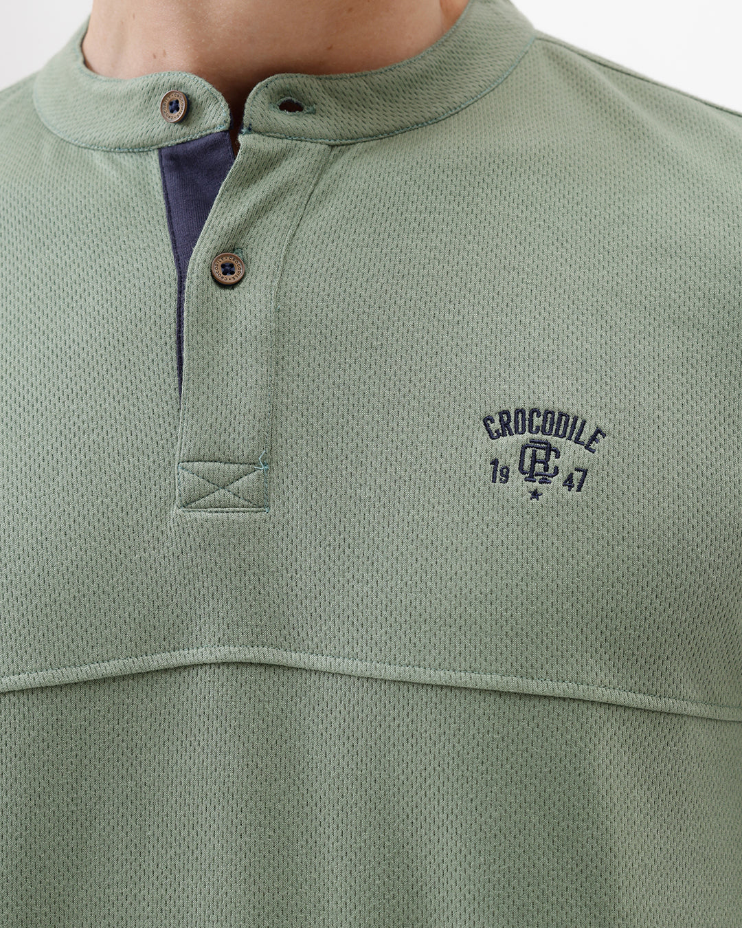 A man wearing a sage green long-sleeve henley shirt with a small embroidered logo on the left chest, paired with blue jeans, against a plain white studio background.