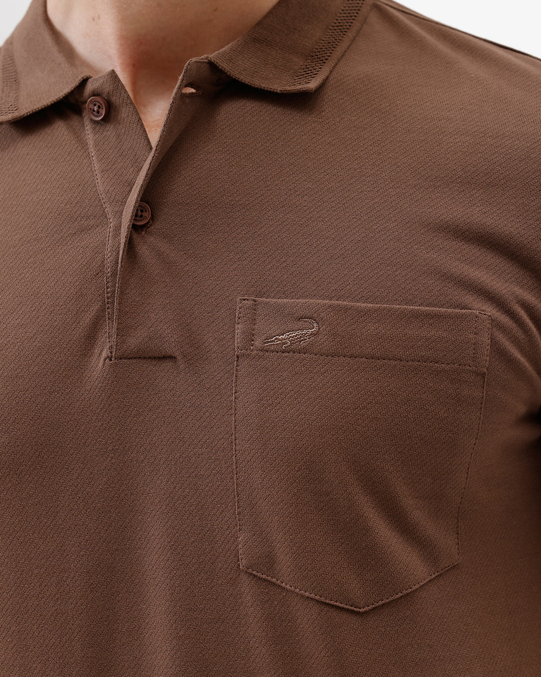A man wearing a brown short-sleeve polo shirt with a chest pocket, paired with blue jeans, standing against a plain light gray background.