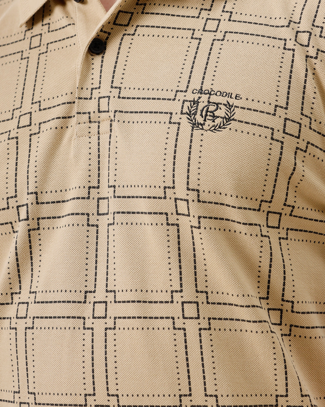 A man wearing a beige short-sleeve polo shirt with a brown grid pattern, paired with black jeans, standing against a plain white studio background.