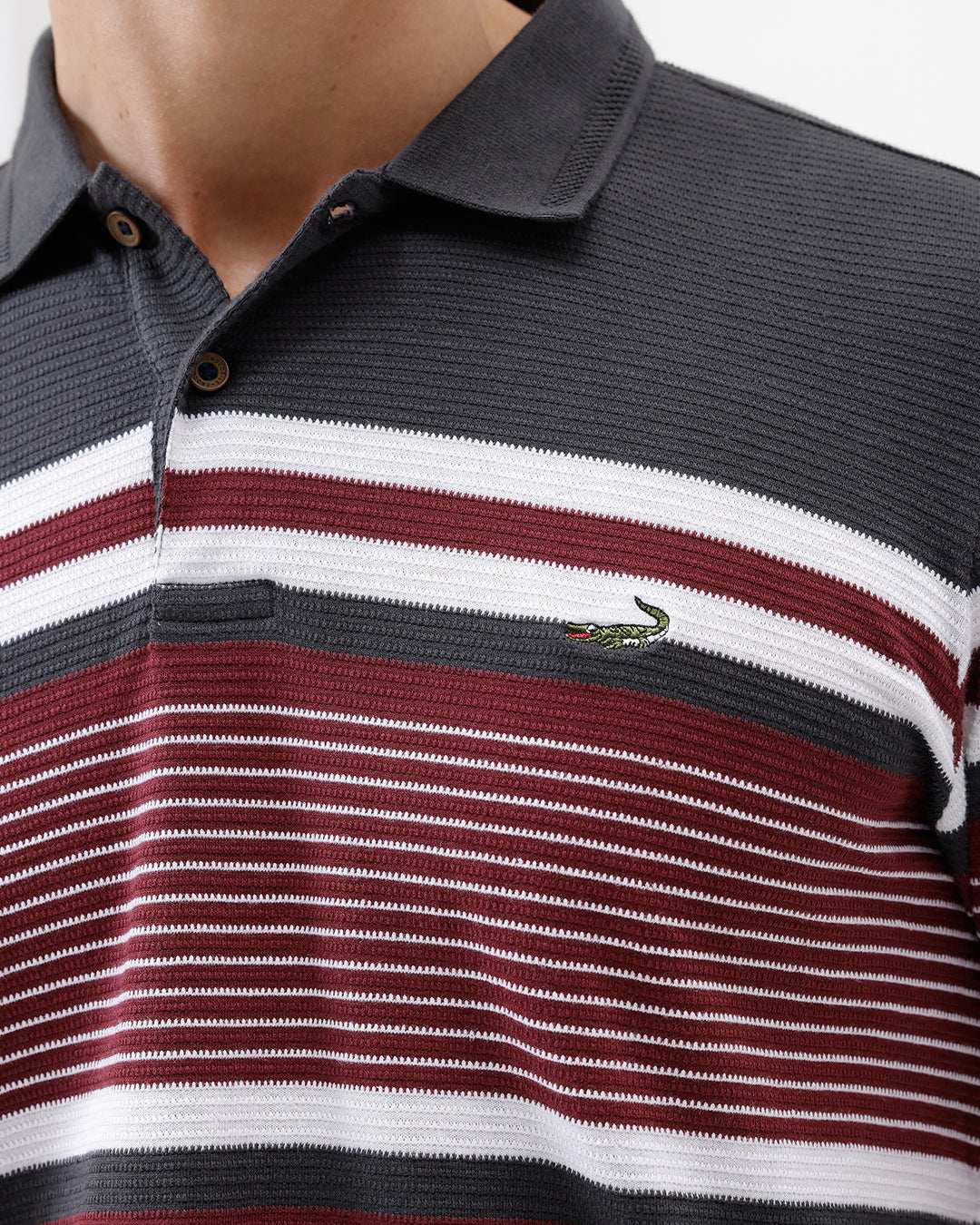 A man wearing a dark gray Lacoste polo shirt with horizontal red and white stripes across the chest and sleeves, paired with blue jeans, against a plain light gray background.