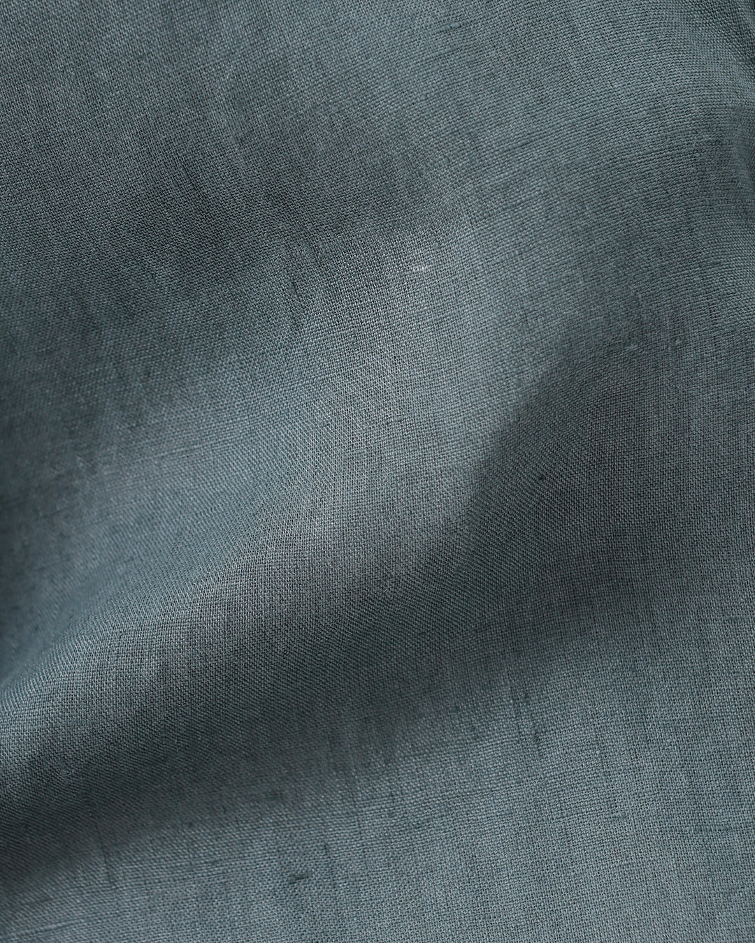 A man wearing a short-sleeve button-up shirt in a muted slate green color, paired with light beige trousers, standing against a plain light gray background.