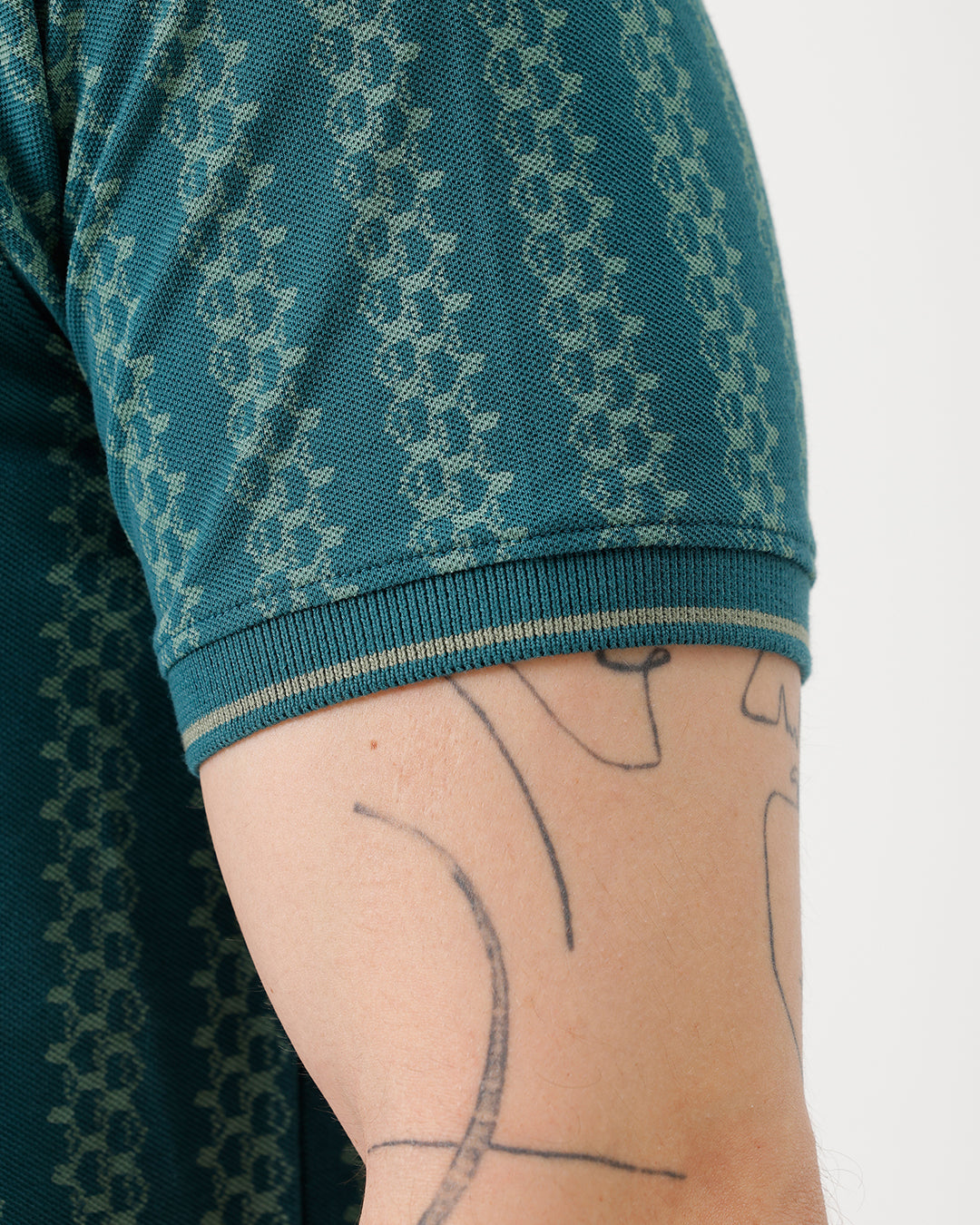 A man wearing a teal green short-sleeve polo shirt with a white geometric pattern, paired with black jeans, standing against a plain white studio background.