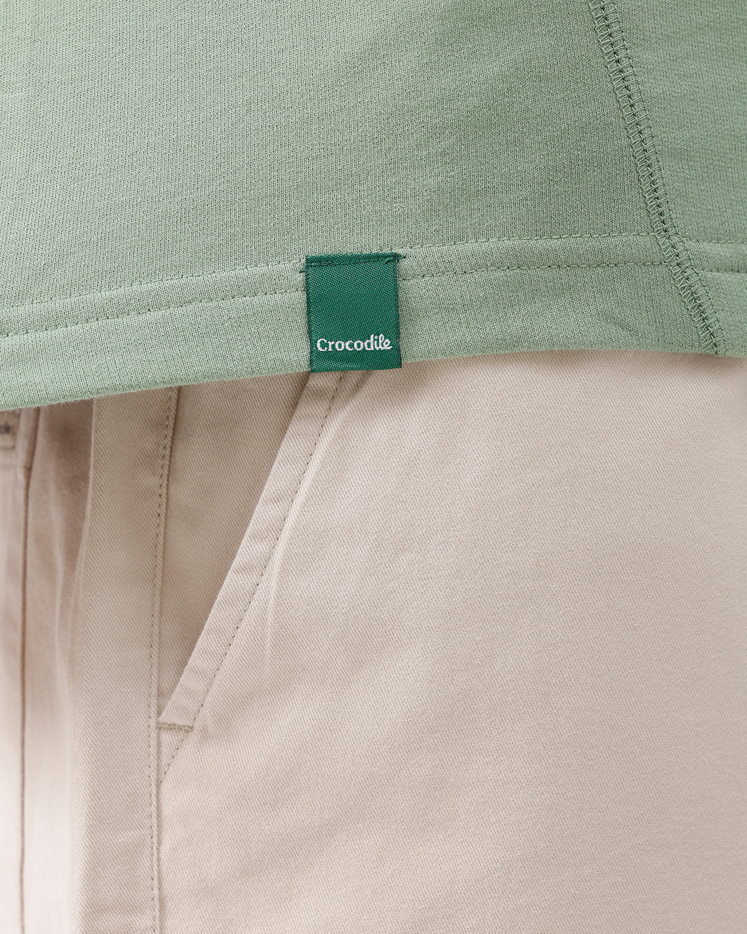 A man wearing a light sage green athletic t-shirt with a small tonal logo on the chest, paired with beige pants, standing against a plain white studio background.