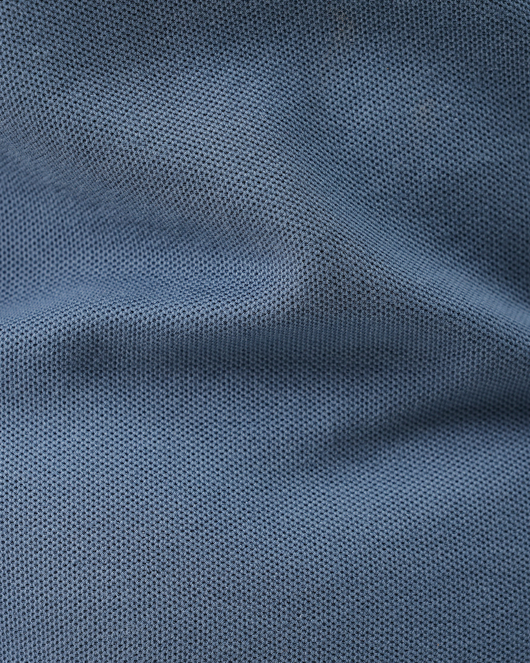 A man wearing a slate blue short-sleeve polo shirt and beige trousers, standing against a plain white studio background.