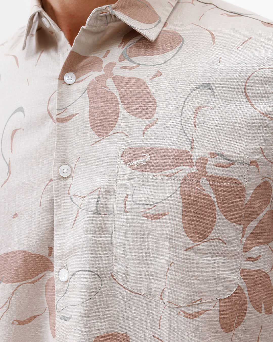 A man wearing a short-sleeve button-up shirt with a beige and brown floral pattern, paired with gray trousers, standing against a plain white background.