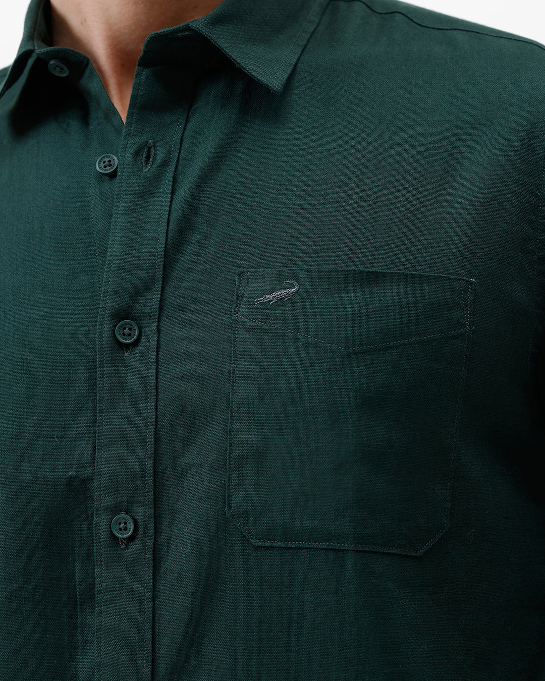 A man wearing a dark forest green long-sleeve button-down shirt with a chest pocket, paired with dark jeans, standing against a plain white studio background.