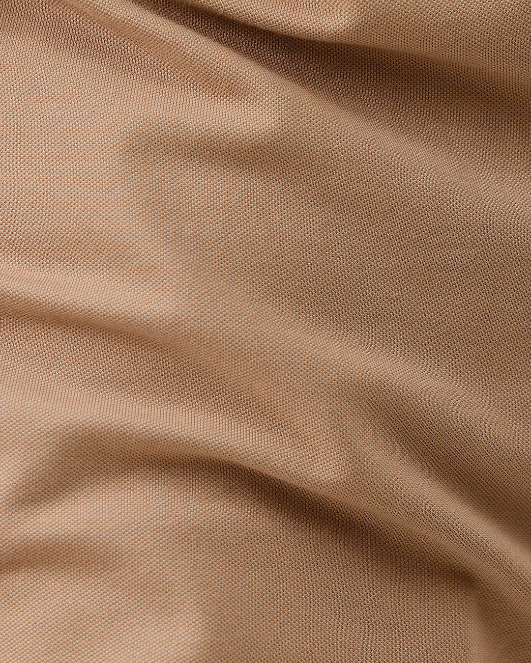 A man wearing a tan short-sleeve polo shirt with dark blue piping on the collar and sleeve cuffs, paired with dark navy pants, against a plain light gray background.