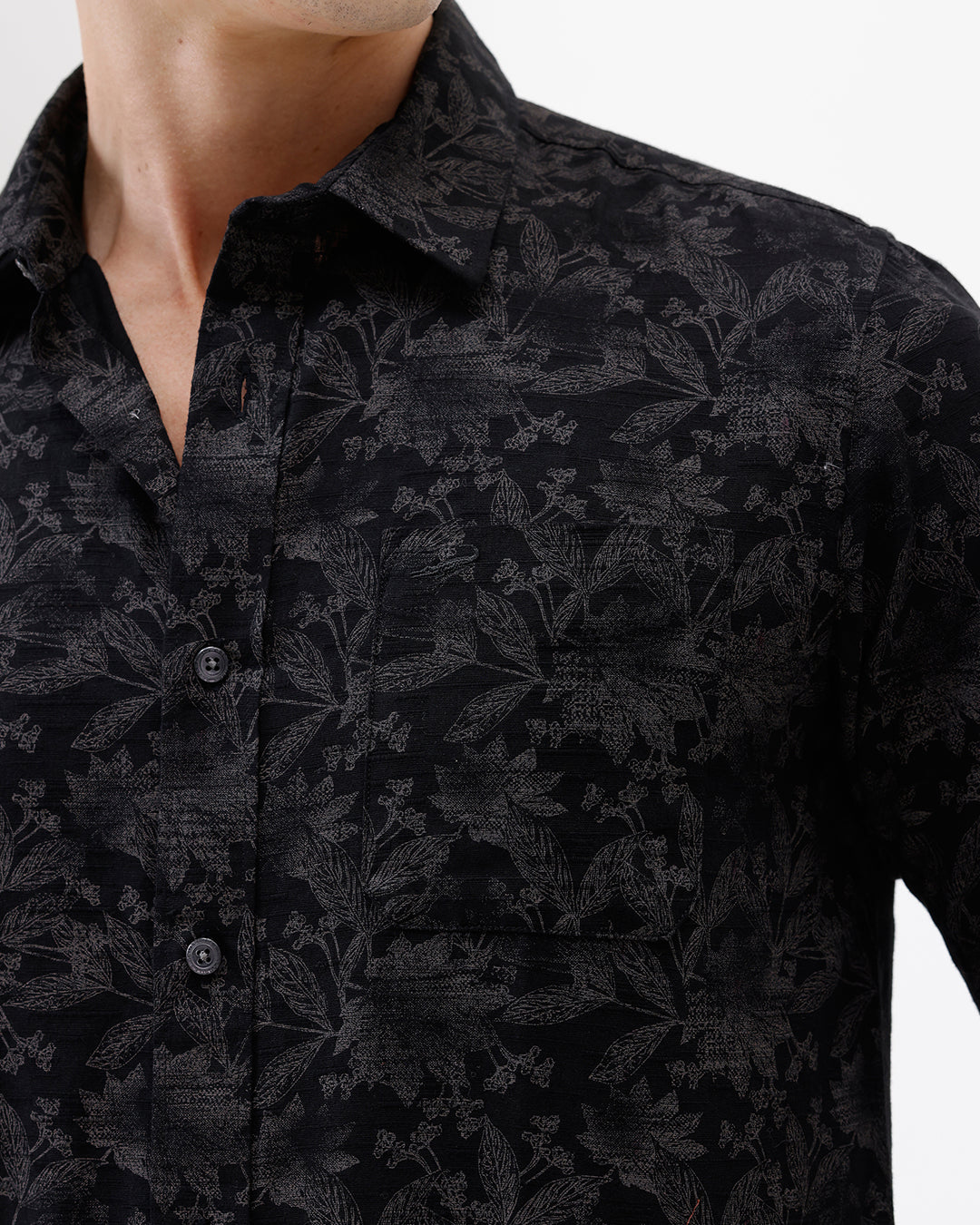 A man wearing a black long-sleeve button-up shirt with a subtle tonal floral pattern, paired with beige cargo pants and dark sunglasses, standing against a plain white background.