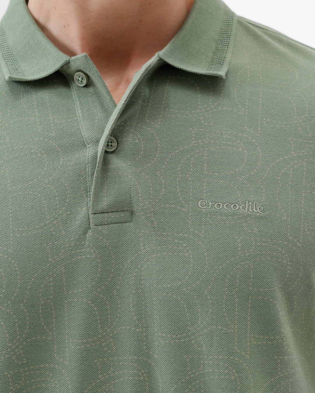 A man wearing a short-sleeve polo shirt in sage green, paired with dark blue jeans, standing against a plain light gray background.