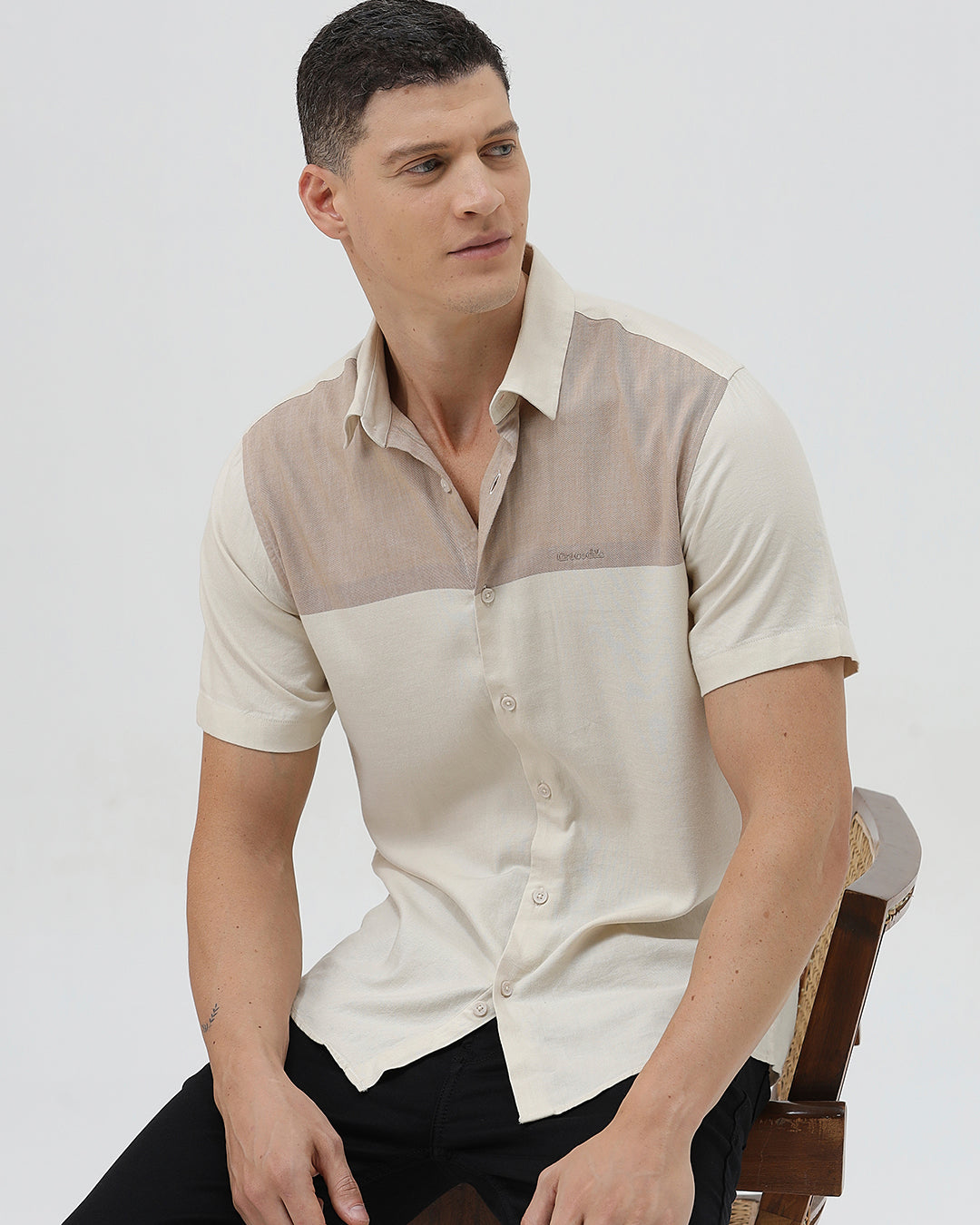 A man wearing a short-sleeve linen button-up shirt with a color-blocked design in beige and cream, paired with black pants, against a plain white studio background.