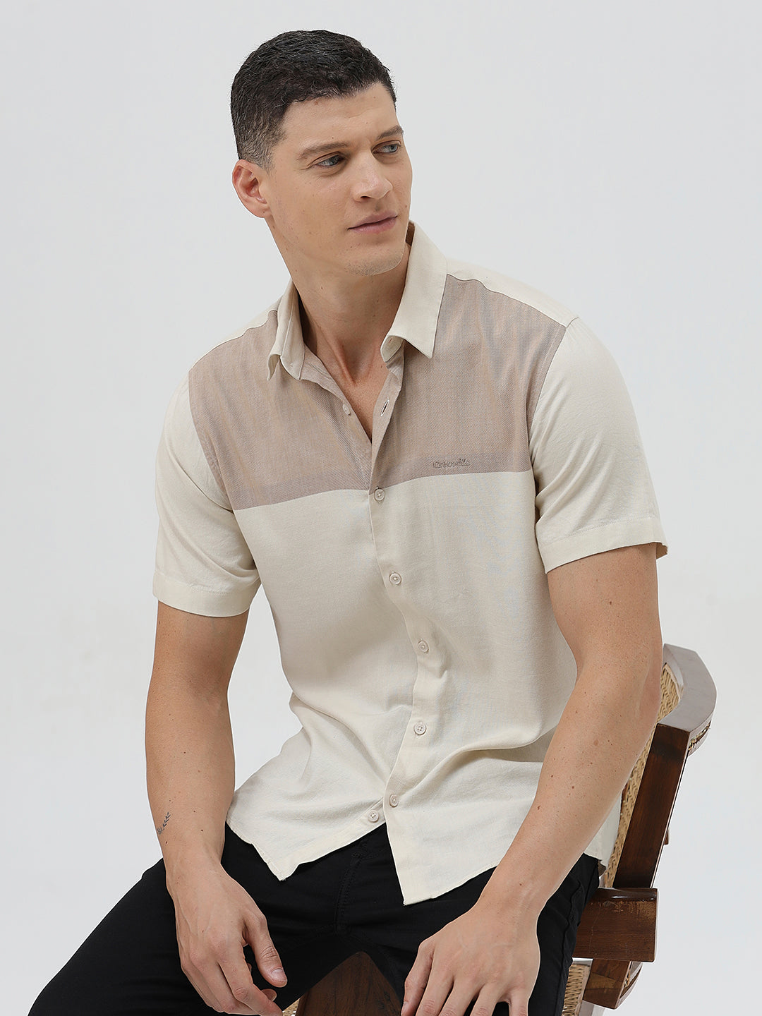 A man wearing a short-sleeve linen button-up shirt with a color-blocked design in beige and cream, paired with black pants, against a plain white studio background.