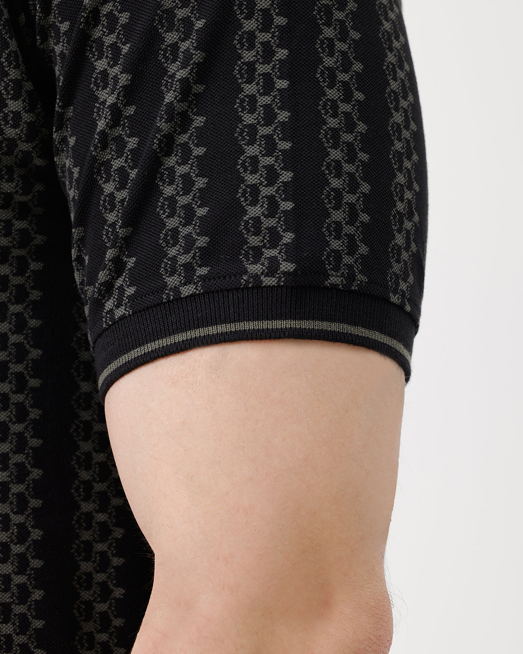 A man wearing a black short-sleeve polo shirt with a subtle geometric pattern, paired with dark gray trousers, standing against a plain white studio background.