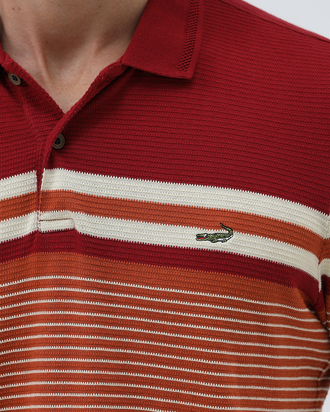 A man wearing a red short-sleeve polo shirt with horizontal cream and rust-orange stripes across the chest and sleeves, paired with dark blue jeans and black sunglasses, against a light gray studio background.