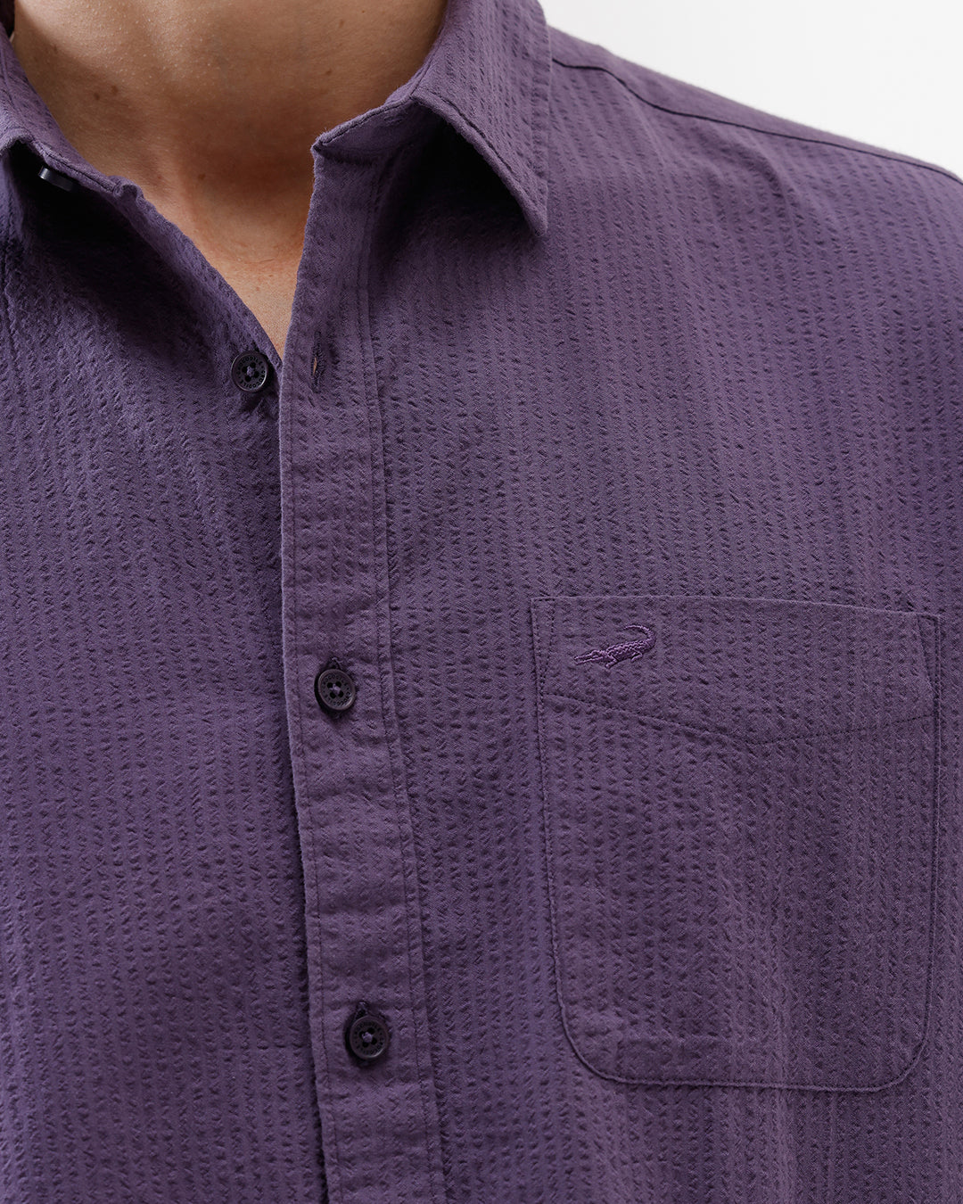 A man wearing a long-sleeve button-down shirt in a muted purple color, paired with black pants, standing against a plain white studio background.