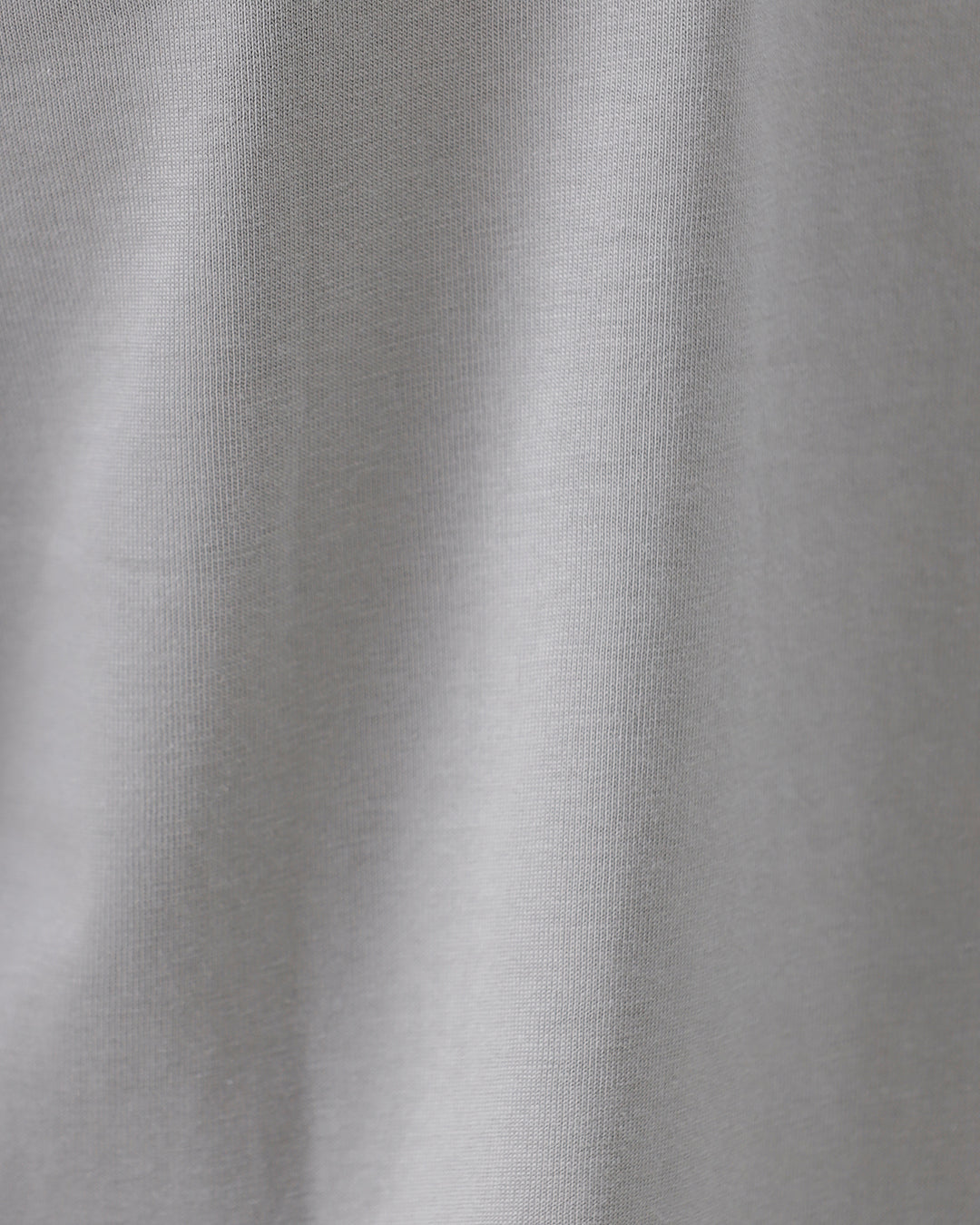 A man wearing a light gray short-sleeve crewneck t-shirt with a subtle tonal chest logo, paired with dark jeans, standing against a light gray studio background.