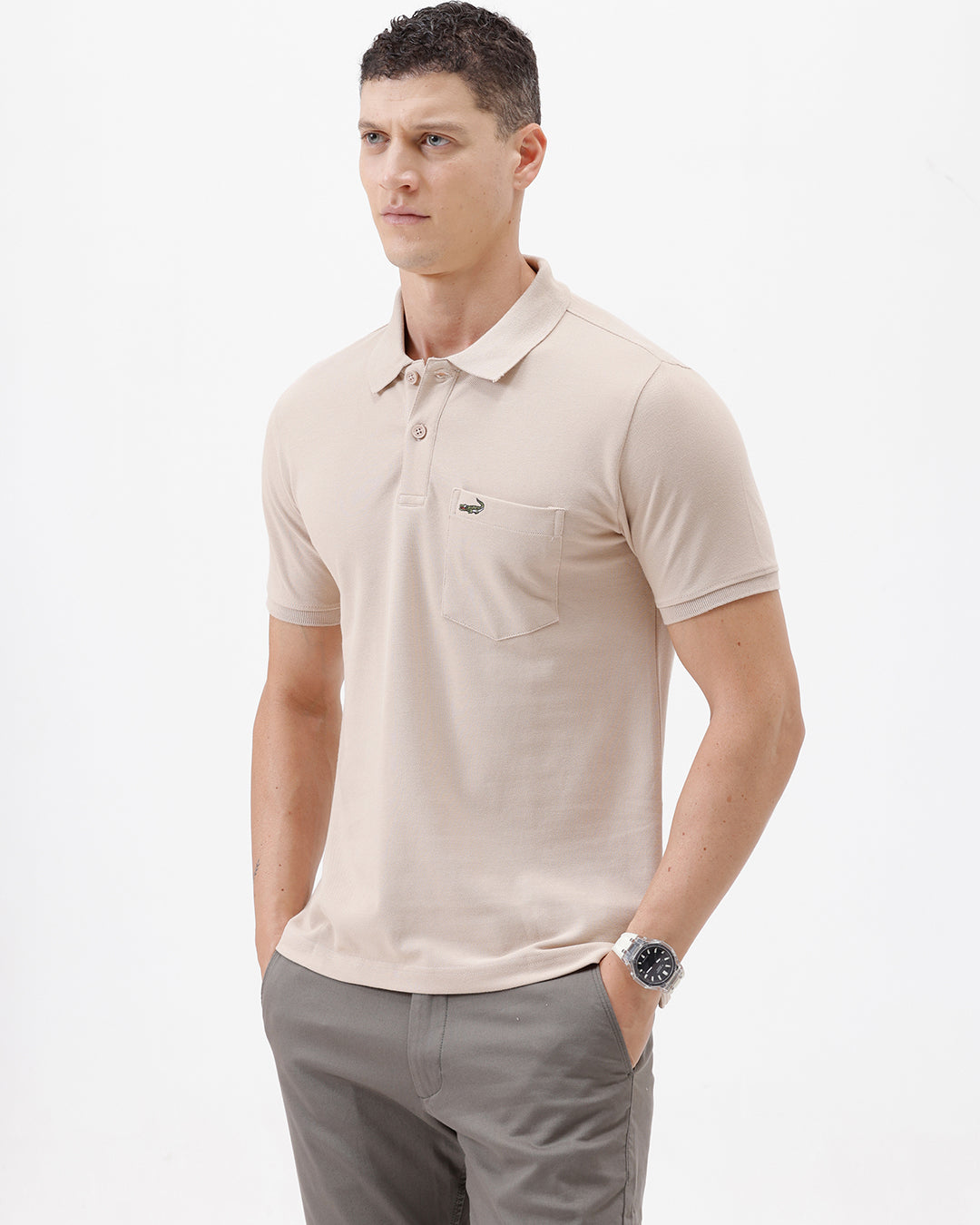 A man wearing a light beige short-sleeve polo shirt with a small crocodile logo on the chest pocket, paired with gray trousers, photographed against a plain white studio background.